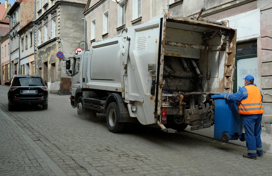 A row of large, plastic wheelie bins lined up along a paved pathway beside a brick wall, with the bins primarily in green, blue, and red colors. The green bin in the foreground has a ridged lid, sligh